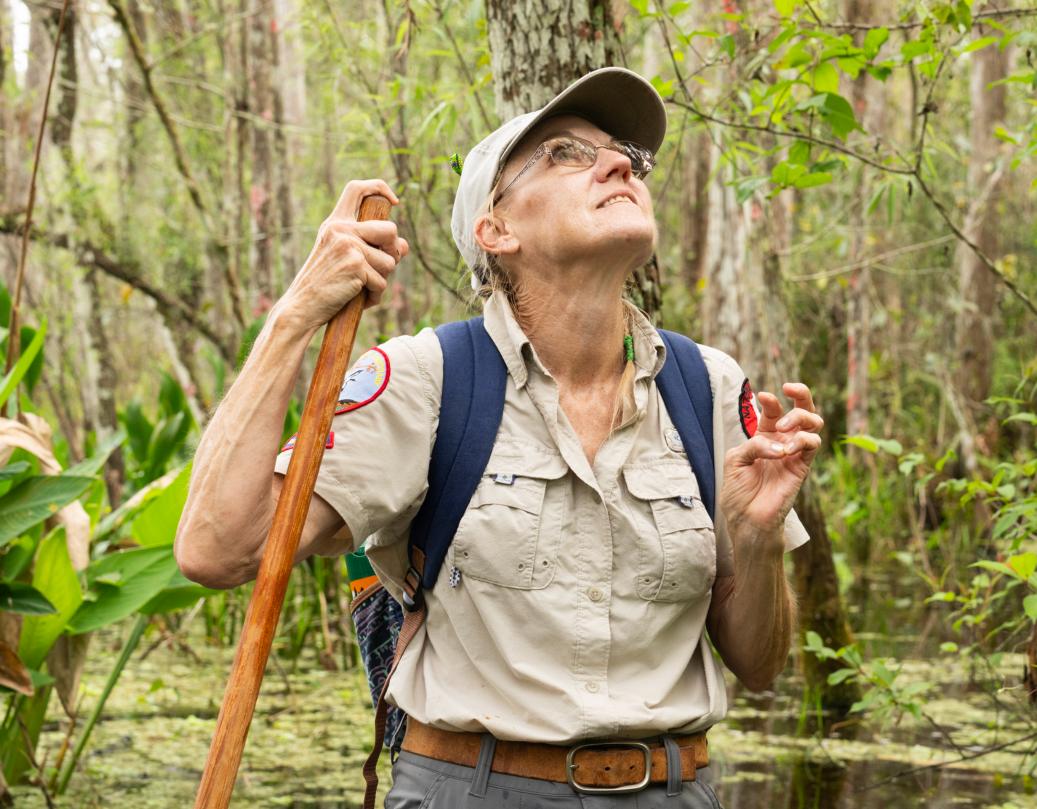 A woman holding a walking stick looks up, with trees in the background